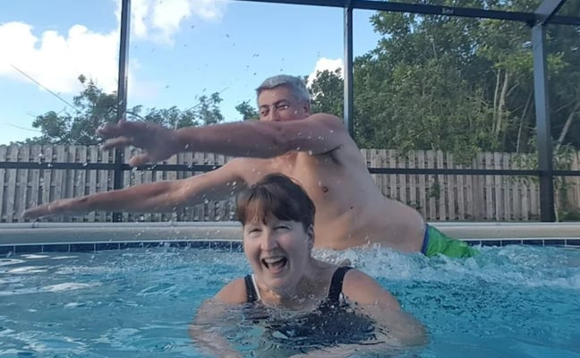 Two people swimming and playing in a screened-in pool on a sunny day with trees visible beyond the fence.